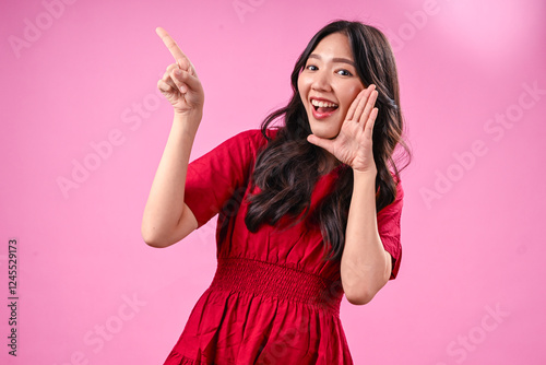 A young Asian woman with long wavy black hair, wearing a red dress, poses against a pink background. She looks excited, whispering with one hand near her mouth while pointing upwards