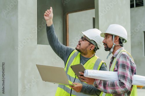 Construction engineers wearing safety helmets and reflective vests analyzing structural elements of unfinished residential project. Digital devices used for on-site plan assessment and coordination.