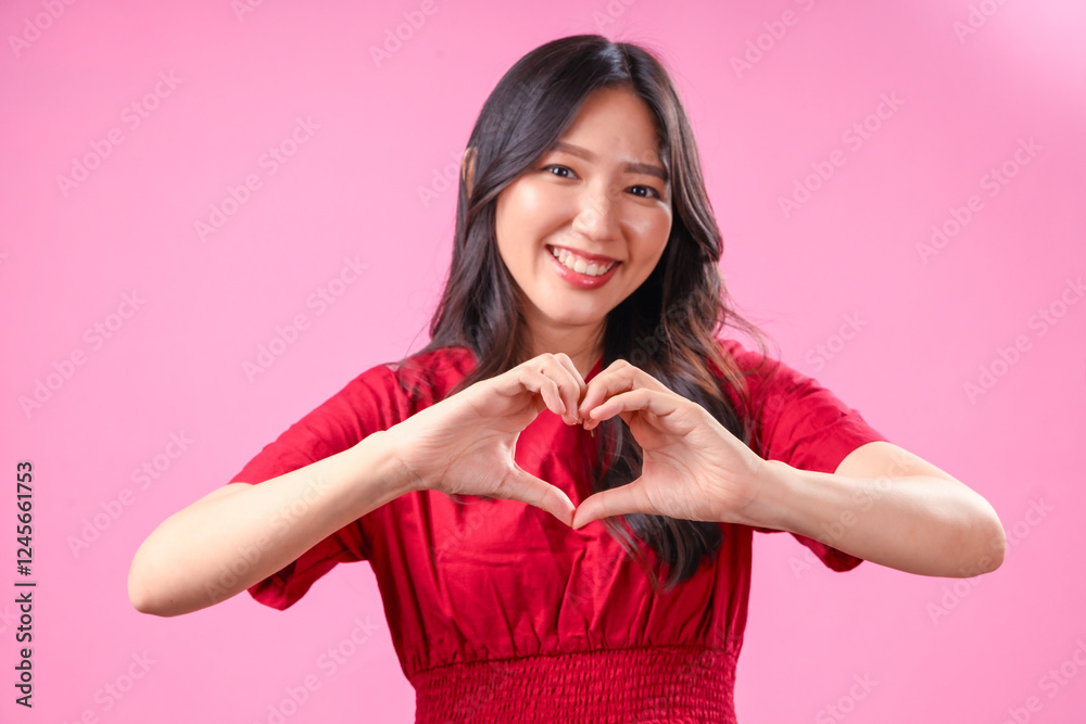 A young Asian woman with long wavy black hair wears a red dress and smiles warmly while forming a heart shape with her hands against a pink background, expressing love, kindness, and positivity A young Asian woman with long wavy black hair wears a red dress and smiles warmly while forming a heart shape with her hands against a pink background, expressing love, kindness, and positivity