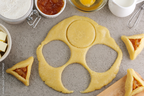 Female hands cutting out a round shape for pastry dough, preparation for the traditional Jewish holiday Purim day. A group of ingredients on the background. top view photo