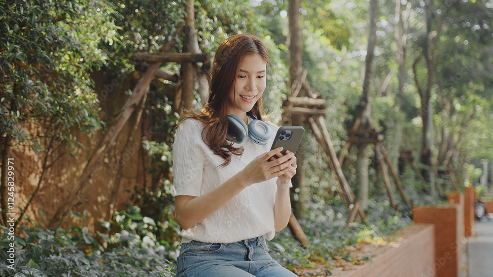 Naklejka premium Attractive young woman enjoys her smartphone while ing headphones in lush outdoor setting, radiating happiness and relaxation. Asian Woman Lifestyle