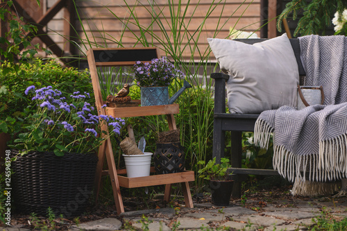 Summer outdoor backyard decorations. Rustic flower stand with potted plants, wooden bench with pillows and blanket. Cozy garden relax area.