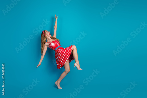 Young woman in a vibrant red polka dot dress against a blue background striking an expressive playful pose