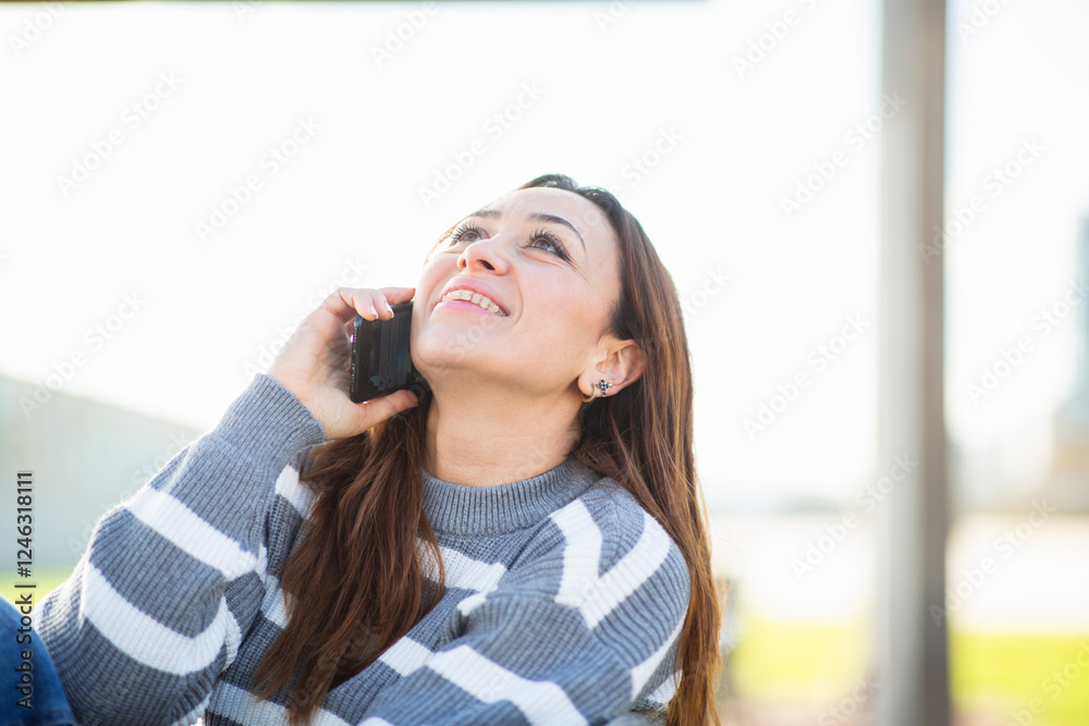 Fototapeta premium Young woman in casual wear talking on smartphone while sitting outdoors