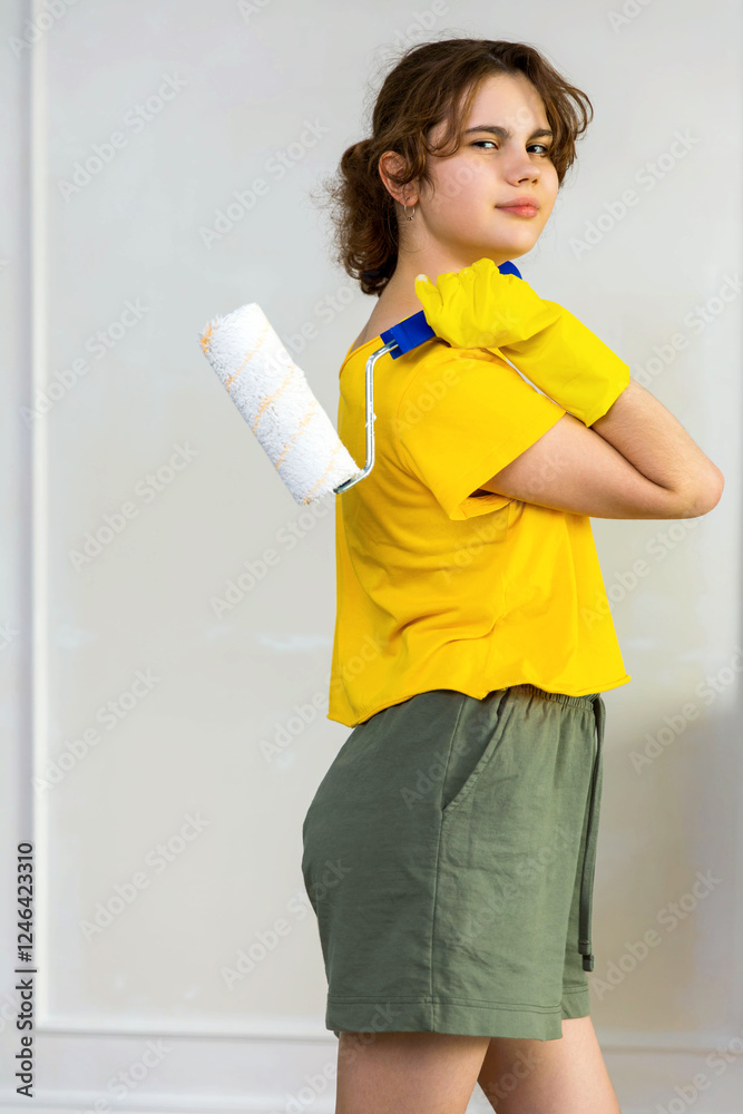 Teenage girl with wide smile, curly haired in yellow t-shirt, yellow rubber gloves holding wall paint roller on wall background. Concept repair, painting is easy and joyful. Teenage girl with wide smile, curly haired in yellow t-shirt, yellow rubber gloves holding wall paint roller on wall background. Concept repair, painting is easy and joyful.