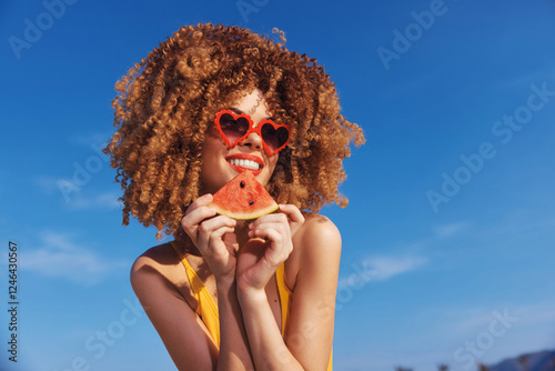 Young woman with curly hair enjoying a slice of watermelon against a bright blue sky She s wearing heart shaped sunglasses photo