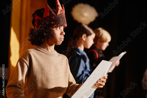 Portrait of young child engaging in school play by wearing medieval costume and holding white prop, while other children are in background participating in same activity