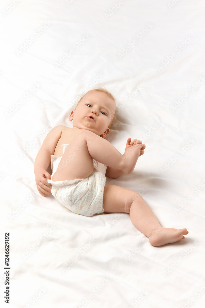 Cute baby, girl in diaper lying in bed and looking at camera with curiosity against white studio background. Vertical image. Concept of childhood, motherhood, care. Copy space for ad Cute baby, girl in diaper lying in bed and looking at camera with curiosity against white studio background. Vertical image. Concept of childhood, motherhood, care. Copy space for ad