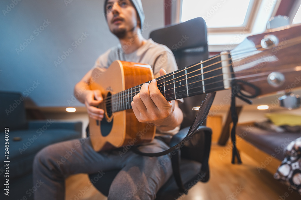 Obraz premium Man alone in the middle of a sunlit room enjoys playing a beautiful wooden acoustic guitar and composing new songs. Immersed in the sweet tones of the guitar. Music that heals the soul