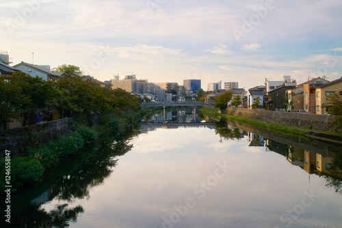 Tranquil river reflecting cityscape and trees under a serene sky in an urban setting, Kanazawa, Japan