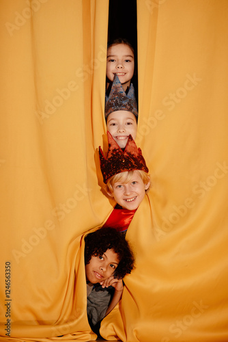 Obraz na plátne Group of children peeking from yellow curtain during holiday performance, smiling and showing enthusiasm for the event