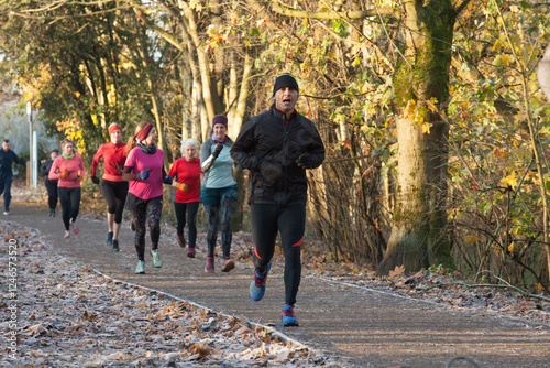 A group of people jogging on a path covered with fallen leaves in a forest, wearing colorful athletic clothing. Sale Water Park, Manchester, UK photo