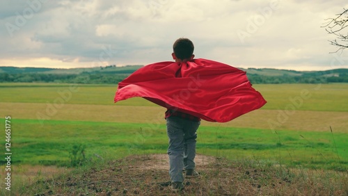 Kid boy in superhero costume runs into distance. Children's party concept. Child superhero red in red cape plays runs in nature. Boy plays superhero red cape. Family walk child dream play in nature photo