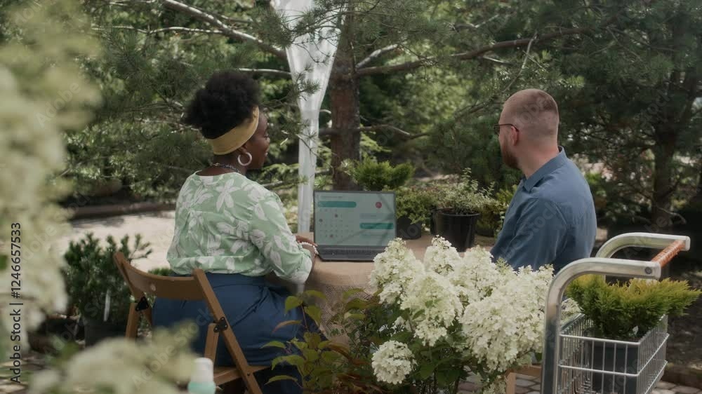 Young Caucasian man and young Black woman in casualwear sitting at table with laptop inside open pavilion at plant nursery among potted tagged shrubs engaged in conversation on summer day Young Caucasian man and young Black woman in casualwear sitting at table with laptop inside open pavilion at plant nursery among potted tagged shrubs engaged in conversation on summer day