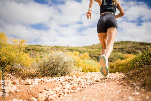 Female trail runner wearing black sportswear and a hydration vest, running through a scenic natural landscape on a rocky path. photo