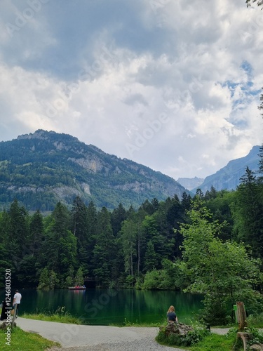 Peaceful scene at Blausee (Blue Lake) in Switzerland, with a mother and child relaxing by the turquoise waters, surrounded by lush green forest and towering mountains under a cloudy sky. photo