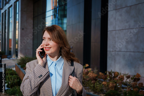 A cheerful young businesswoman in a stylish blazer is having a phone conversation outdoors in an urban setting. She is smiling confidently, exuding positivity and professionalism. 