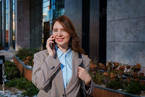 A cheerful young businesswoman in a stylish blazer is having a phone conversation outdoors in an urban setting. She is smiling confidently, exuding positivity and professionalism. 