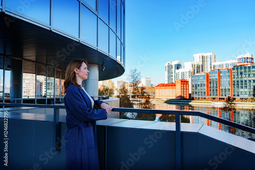 A young woman in a blue coat stands on a terrace, gazing at the modern city skyline with a calm and thoughtful expression. The glass building behind her reflects the urban landscape.