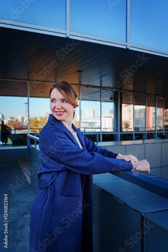 A stylish young woman in a blue coat stands outside a modern glass building, leaning on a railing and smiling. The reflective windows of the structure capture the surrounding cityscape.