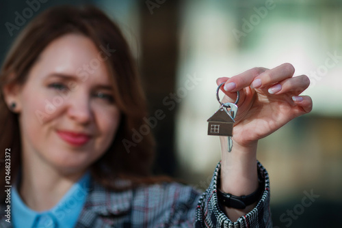 Smiling woman holding house keys with a home-shaped keychain. Represents real estate, home purchase, renting, or investment. Modern city background, professional and confident look.