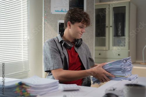 Young student is sitting at a desk, organizing a large stack of papers. Headphones rest around his neck, suggesting a break from studying or working on a project