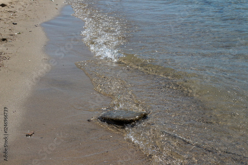 Fototapeta Naklejka Na Ścianę i Meble -  Summer vacation, calm, clean sea water, the view by the sea with waves. Brown yellow sea sand on the beach, bright sun reflecting on big and small sea stones. Balikesir, sarimsakli, Türkiye