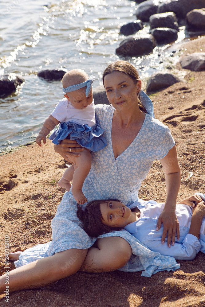 A happy mother in a dress with two children aged seven and a baby are standing on a sandy beach by the sea in summer