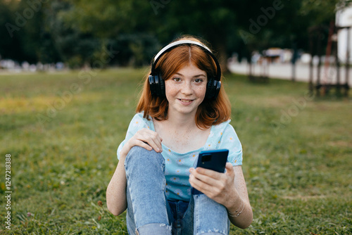 Redhaired teenage girl with freckles in wireless headphones and smartphone sitting on the grass