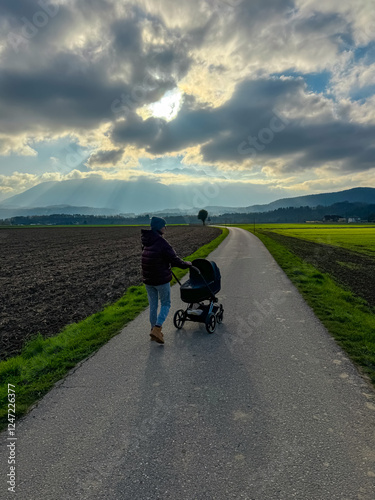 Parent pushing stroller on rural path at sunset in St. Egyden, Austria. Dramatic cloudy sky and Karawanks mountain range in background. Plowed field and green meadow frame peaceful countryside scene photo