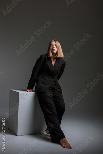 Fotografía Stylish woman in a black suit poses on a white cube in a minimalist studio setti
