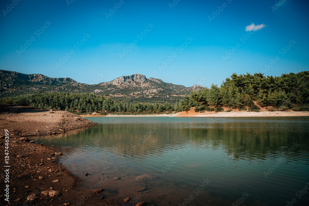 A serene mountain lake surrounded by lush pine forests and rugged hills under a vibrant blue sky. The calm water reflects the natural beauty of the landscape, creating a picturesque view A serene mountain lake surrounded by lush pine forests and rugged hills under a vibrant blue sky. The calm water reflects the natural beauty of the landscape, creating a picturesque view