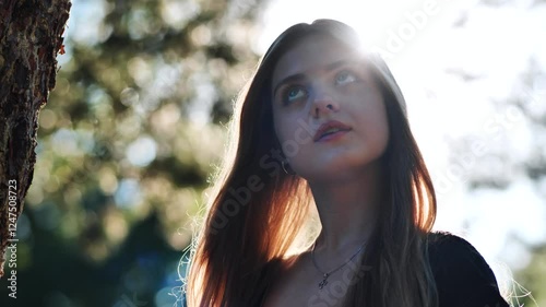 Female graduate contemplating future, standing amid sunlit woodland, golden backlight illuminating pensive moment
