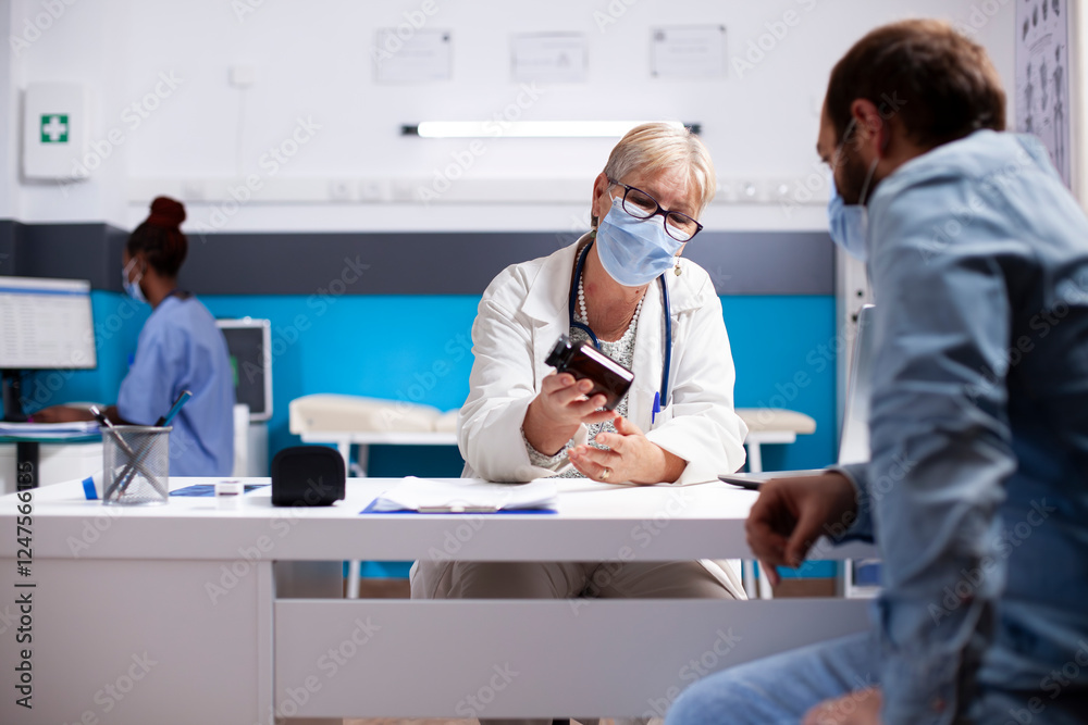 Fototapeta premium Caucasian young man listens attentively as senior doctor explains treatment, holding pills bottle in medical office. Older female medic describes usage of medicine to patient for successful recovery.
