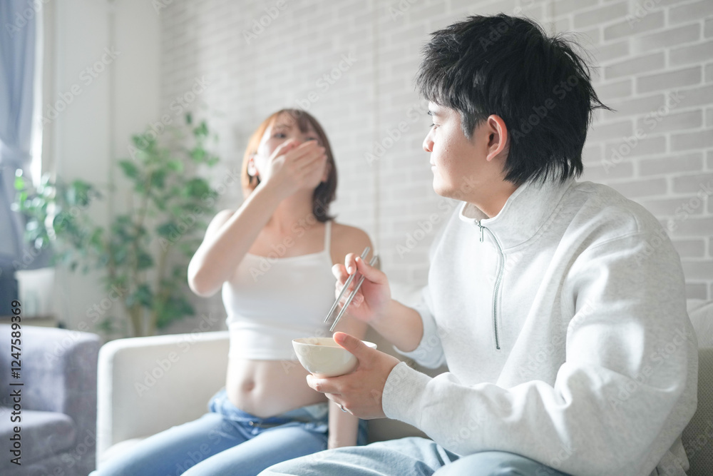 A Japanese pregnant woman in her 20s, wearing a dress that shows her stomach, sits on a sofa in an apartment and her husband eats a hot pot dish. A Japanese pregnant woman in her 20s, wearing a dress that shows her stomach, sits on a sofa in an apartment and her husband eats a hot pot dish.