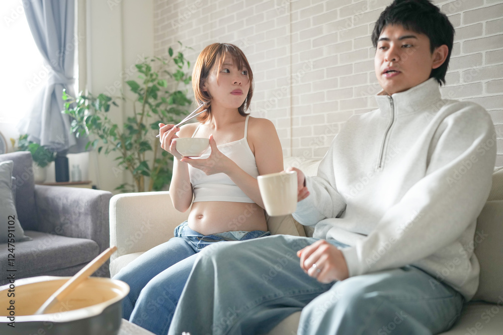 A Japanese pregnant woman in her 20s, wearing a dress that shows her stomach, sits on a sofa in an apartment and her husband eats a hot pot dish. A Japanese pregnant woman in her 20s, wearing a dress that shows her stomach, sits on a sofa in an apartment and her husband eats a hot pot dish.