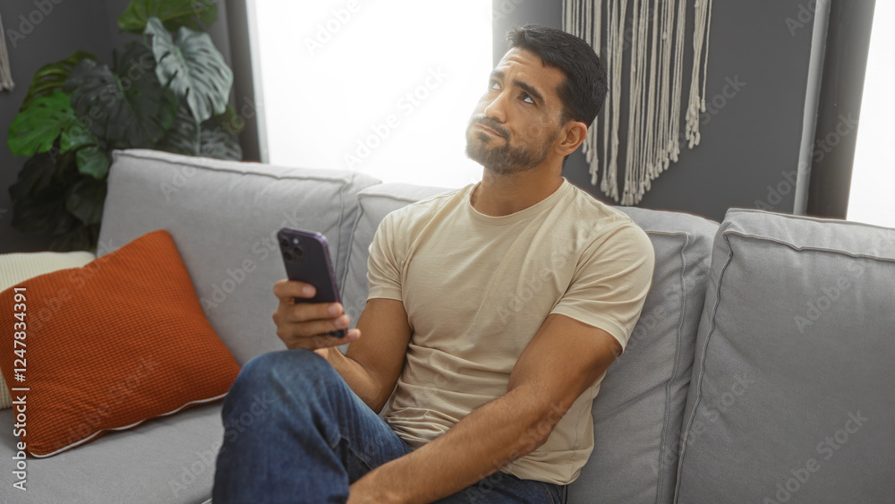Obraz premium Young man with beard relaxing in living room holding phone, sitting on sofa, with pillow and plants, looking thoughtful, representing home comfort, modern interior design.