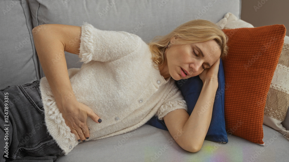 Woman resting on comfortable sofa in bright living room showing signs of discomfort with hand on stomach, beautiful blonde middle-aged adult relaxing indoors. Woman resting on comfortable sofa in bright living room showing signs of discomfort with hand on stomach, beautiful blonde middle-aged adult relaxing indoors.
