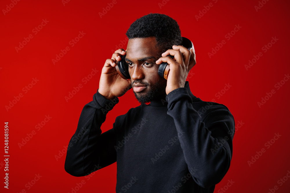 Young Black man wearing headphones against a vibrant red background, focused and enjoying music, capturing emotions of relaxation and engagement Young Black man wearing headphones against a vibrant red background, focused and enjoying music, capturing emotions of relaxation and engagement