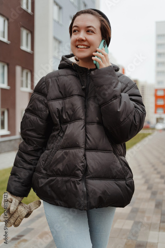 A beautiful young woman enjoys a winter day, walking through a snowy city and chatting on her phone. She laughs and smiles brightly, exuding happiness and a cozy lifestyle vibe in the urban setting