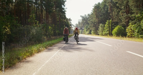 A man and a woman ride bicycles on an asphalt road surrounded by forest. A sunny day