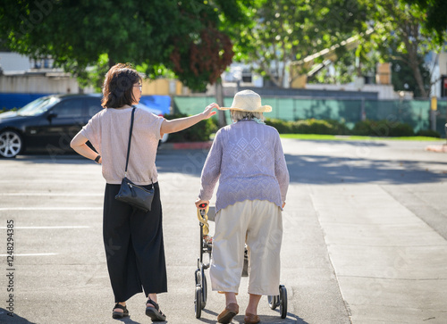 Senior woman walking using a mobility walker in the carpark. Adult daughter adjusting elderly mother’s hat. California.