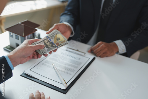 Two businessmen exchanging money and signing a contract over a white table with a miniature house on it, representing a real estate transaction