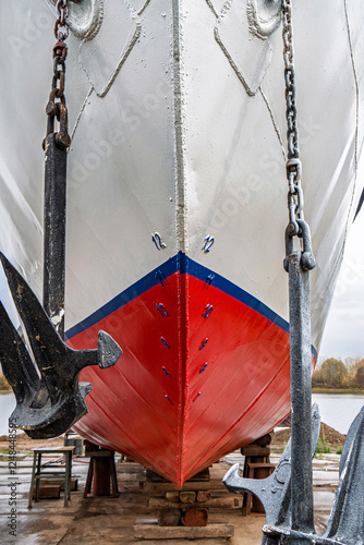 Ship's bow with a red and white hull, flanked by two large anchors on chains close-up. Vessel is docked on dry land for maintenance