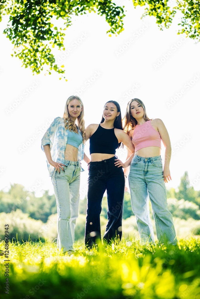 Fototapeta premium Female students smiling, standing close together in lush park setting, sharing summer friendship moment under bright sunlight