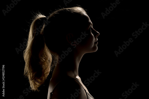 Silhouette of a woman with blonde ponytail, head tilted back. Dramatic side lighting highlights facial features and neck. Dark background crates high contrast.