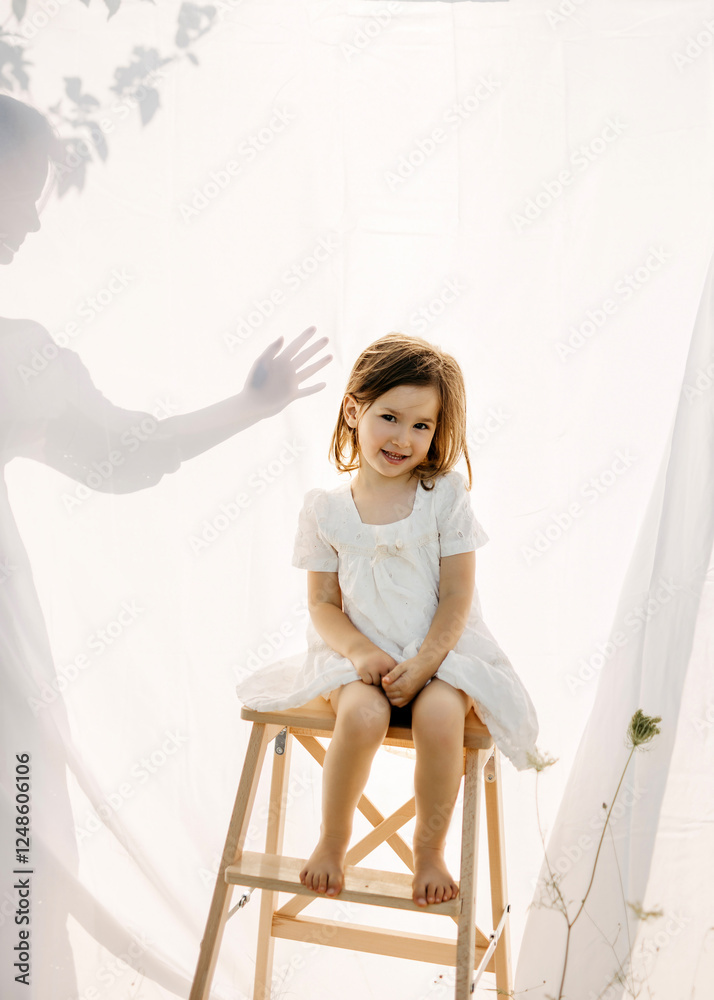 Little happy girl in white dress sits on a wooden ladder, next to her mother shadow reaching hand on background. Little happy girl in white dress sits on a wooden ladder, next to her mother shadow reaching hand on background.
