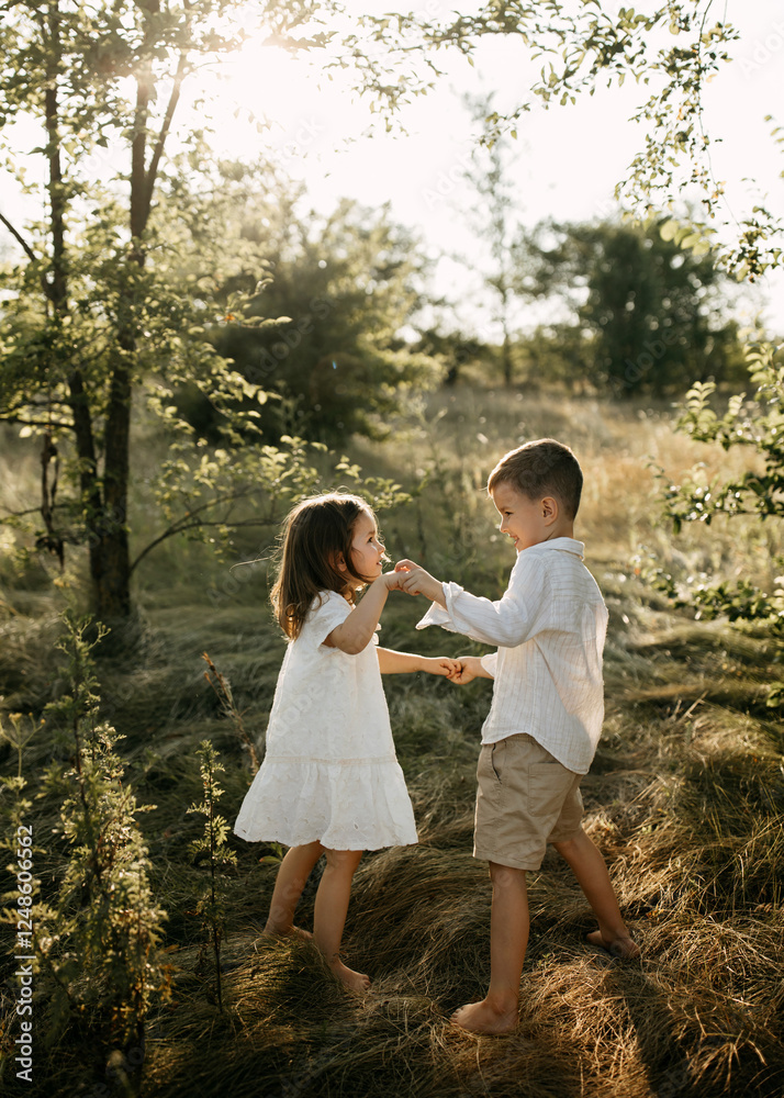 Fototapeta premium Two children holding hands and playing in a sunlit field. Brother and sister playing outdoors on summer day, barefoot.