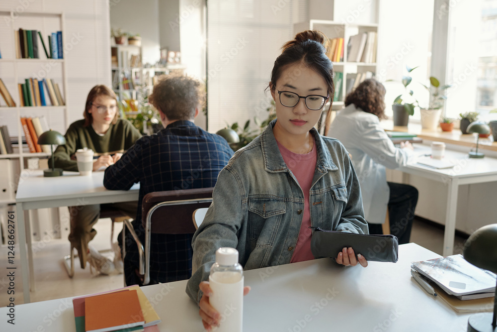 Obraz premium Candid shot of Asian young woman at desk settling in for study session in school or college library with students in background copy space 