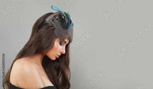 Young female model with healthy long wavy hairstyle, trendy make-up and hairdeco feathers accessories on her head looking down, fashion beauty portrait photo